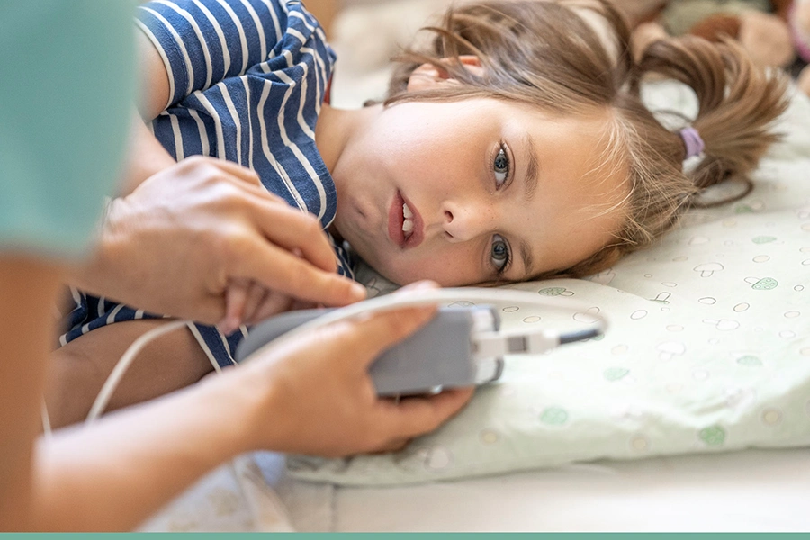 Kind liegt seitlich im Bett mit Blick auf ein Gerät und dabei fast in die Kamera. Das Gerät ist in der Hand einer Pflegerin, die man nur sehr angeschnitten sieht.