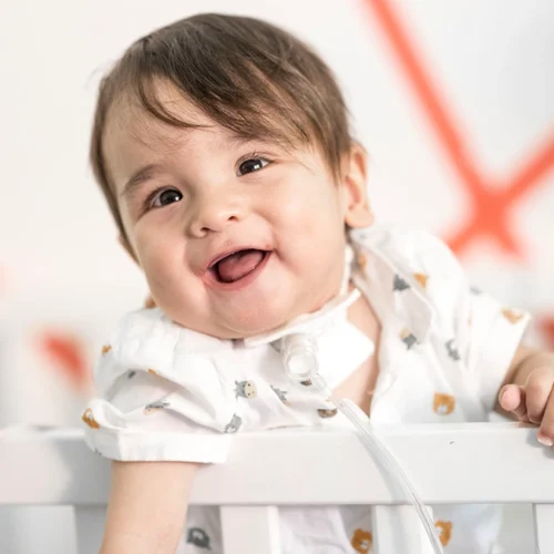 Kleinkind steht im Kinderbett, hält sich am Bettgeländer fest und lacht herzhaft in Richtung Kamera.