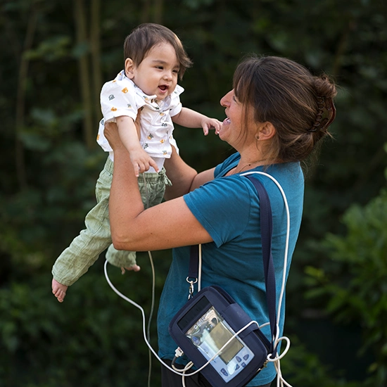 kindl-blogbeitrag-02 Weibliche Pflegekraft mit mit einem Kleinkind im Arm in die Luft hebend. Im Hintergrund ist Wald verschwommen zu sehen.