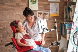 kindl-blogbeitrag-03 Kind im Rollstuhl schaut auf einen Monitor, der dort befestigt ist. Eine Pflegekraft steht daneben und schaut ebenso lächelnd auf den Bildschirm.